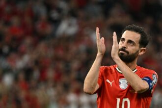 Soccer player in a red jersey with number 10 clapping his hands on the field, blurred stadium in the background.