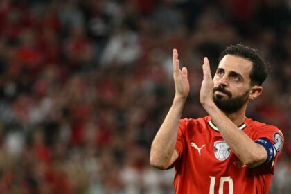Soccer player in a red jersey with number 10 clapping his hands on the field, blurred stadium in the background.