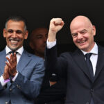Two men in dark suits smiling and clapping, one raising a clenched fist in celebration against a dark background.