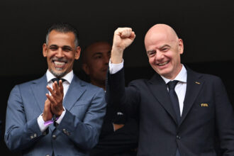 Two men in dark suits smiling and clapping, one raising a clenched fist in celebration against a dark background.