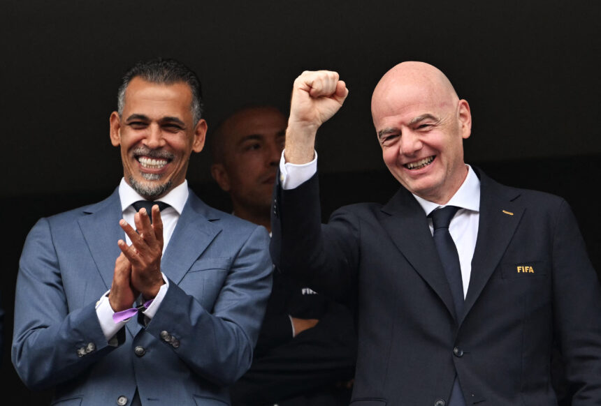 Two men in dark suits smiling and clapping, one raising a clenched fist in celebration against a dark background.