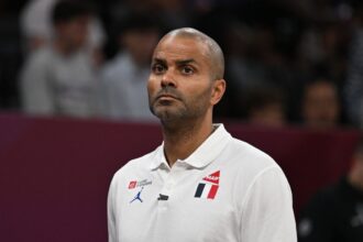 Portrait of a man with a shaved head wearing a white polo shirt with logos, looking upward in a stadium setting