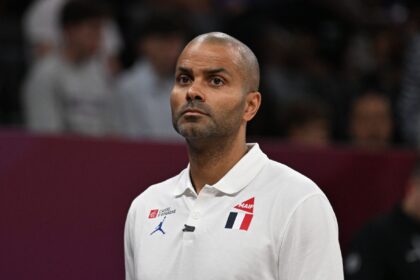 Portrait of a man with a shaved head wearing a white polo shirt with logos, looking upward in a stadium setting