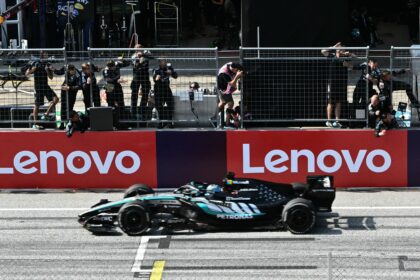 Formula 1 car speeds along the track past the pit wall, with team members watching behind a safety fence and Lenovo banners in the background.