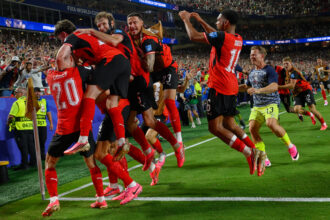 Soccer players in red and black celebrate a goal near the sideline as fans cheer in a packed stadium.