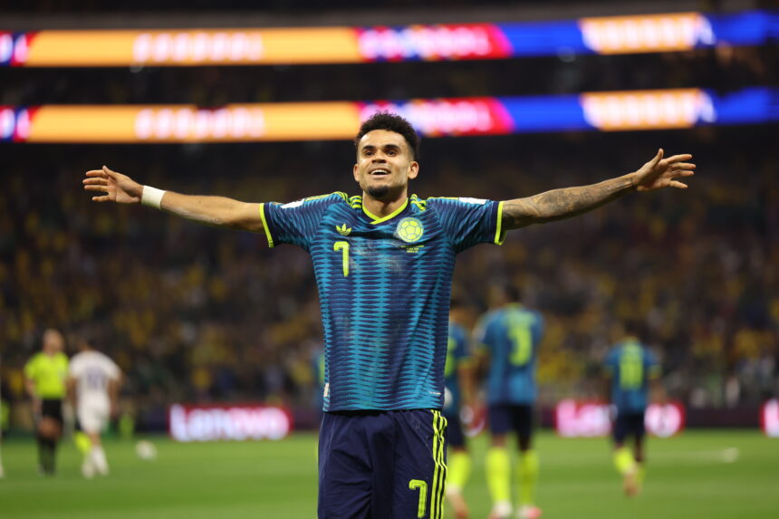 Soccer player in a blue Colombia kit stands with arms outstretched in celebration on the field, a cheering stadium in the background.