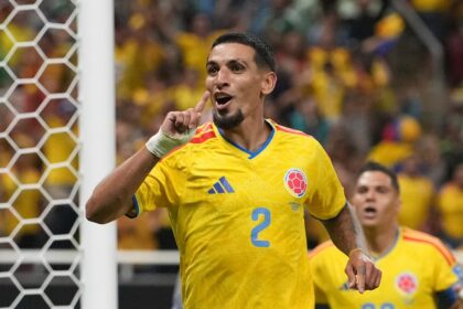 Colombian footballer in a yellow jersey with number 2, celebrating a goal by pointing his finger, near the net with a crowded stadium in the background.