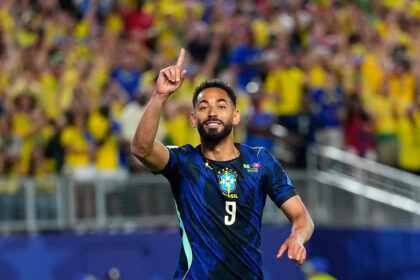 Brazilian soccer player in a blue jersey with raised index finger, celebrating as a sea of yellow-clad fans cheers behind him.