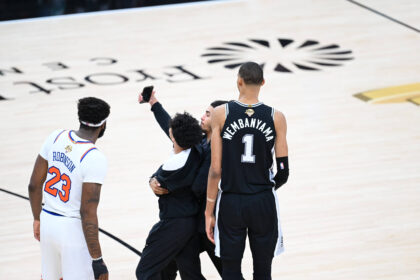 Basketball players on a court, one in a black San Antonio-style jersey with the name Wembanyama and number 1, facing away from the camera alongside teammates in white uniforms; a person in black holds up a phone.