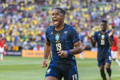 Brazilian soccer player wearing #19 in blue celebrating a goal on the field, stadium crowd in yellow in the background.