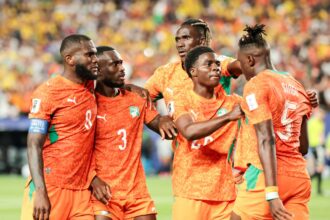 Soccer players in orange jerseys celebrate together on the field with teammates' arms around each other.