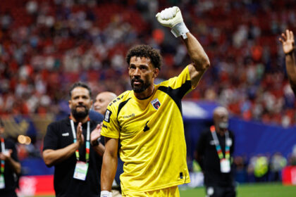 Soccer goalkeeper in a yellow kit raises a gloved fist in celebration on the field with cheering crowd behind him