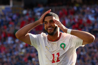 Moroccan footballer celebrating, hands on head, wearing white jersey with green-red Morocco crest, blurred stadium crowd in the background.