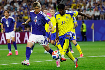Two soccer players, one in blue (Japan) and one in yellow (Sweden), chase the ball on a green field with a crowded stadium in the background.