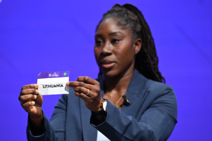 Female presenter holding a Lithuania card up to the camera at a stage event against a blue backdrop.