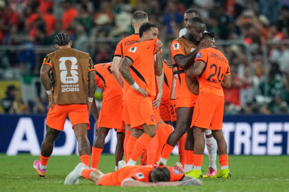 Group of soccer players in bright orange kits celebrate on the field, one player lying on the ground while teammates hug and congratulate him.