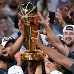 Basketball players lift a gold NBA Championship trophy amid cheering teammates and fans.