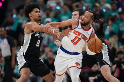 Two basketball players clash for the ball: Knicks player in white #11 grips the ball while Spurs defender in black reaches in, with a crowded arena in the background.
