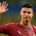 Portugal footballer in a maroon jersey raises his left hand in a wave during a match with a crest visible on the chest.