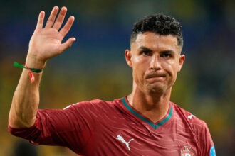 Portugal footballer in a maroon jersey raises his left hand in a wave during a match with a crest visible on the chest.