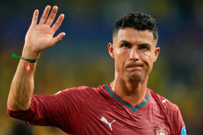 Portugal footballer in a maroon jersey raises his left hand in a wave during a match with a crest visible on the chest.