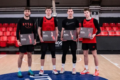 Four young male basketball players stand on a court, each holding a black box/award with 'Ačiū' text, red jerseys and black uniforms, red arena seats in the background.