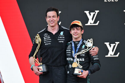 Two race drivers on a podium pose with gold trophies, one older and one younger, in racing suits with sponsor logos.