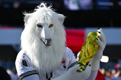 White llama mascot in a white jersey, holding a shiny gold trophy at a stadium event.