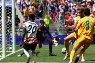 Soccer goal area with players contesting the ball; defender wearing white jersey #20 Balogun faces two yellow-shirted attackers as crowd watches.