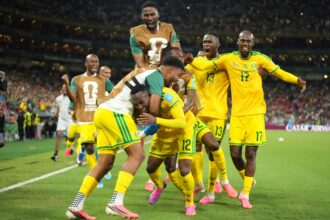 Soccer players in yellow uniforms celebrate together on the field, arms raised, in a packed stadium at nightfall.
