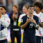 Two male athletes applaud on a track, one in a white USA jacket and the other in navy holding a water bottle, blurred crowd behind them.
