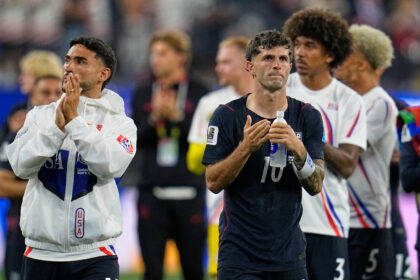 Two male athletes applaud on a track, one in a white USA jacket and the other in navy holding a water bottle, blurred crowd behind them.