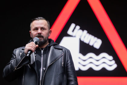 Man in a black leather jacket holding a microphone on stage with a bold red triangular backdrop behind him.