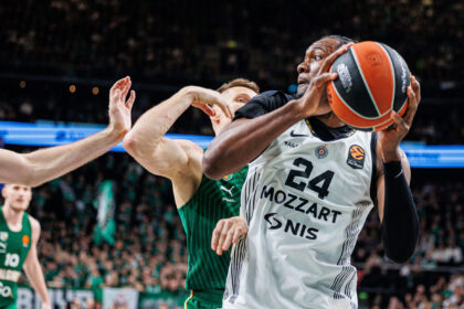 Basketball player in white jersey (no. 24) grips the ball as a green-jersey defender reaches in during a crowded game.