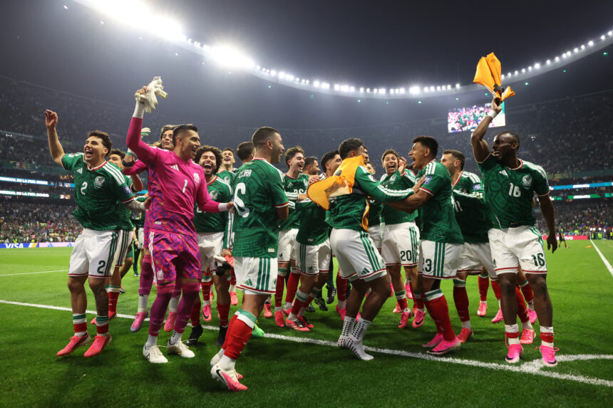 Mexican national team players celebrate on the pitch, hugging and lifting orange training vests amid bright stadium lights.”