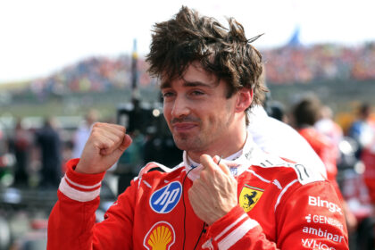 Male race car driver in a red Ferrari suit celebrating with fists raised at a crowded event at the track.