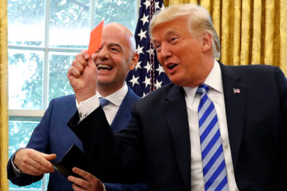 Two men in suits smiling and holding a small orange card in a room with an American flag and gold curtains behind them.