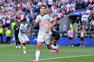 Soccer player in a light-blue patterned jersey runs on the field with a cheering crowd behind him.