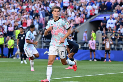 Soccer player in a light-blue patterned jersey runs on the field with a cheering crowd behind him.