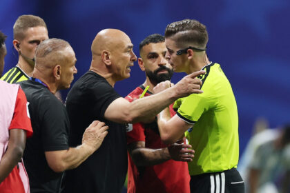 Tense moment on a soccer field as players in red and black confront a teammate in a neon-yellow jersey, hands raised