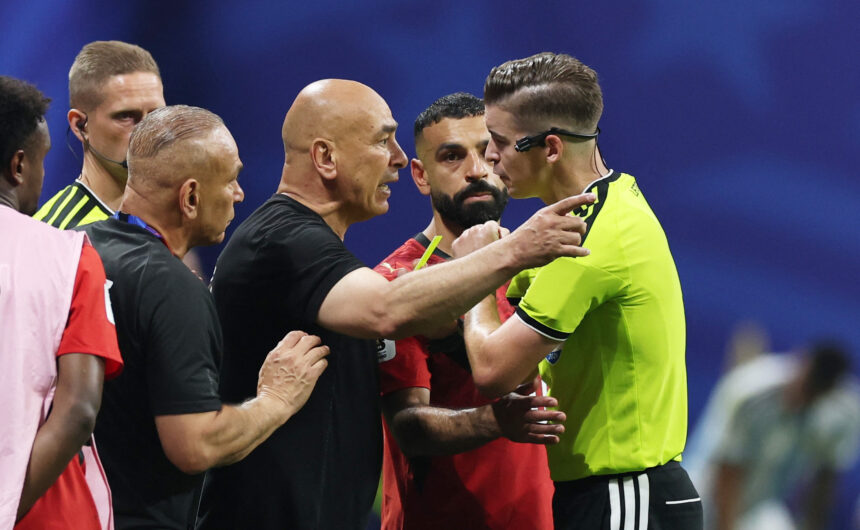 Tense moment on a soccer field as players in red and black confront a teammate in a neon-yellow jersey, hands raised