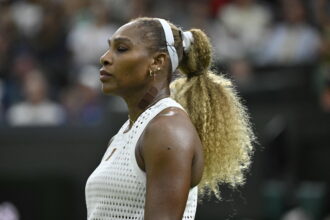 Profile view of a female tennis player wearing a white perforated sleeveless top and a white headband during a match, with a blurred crowd in the background.