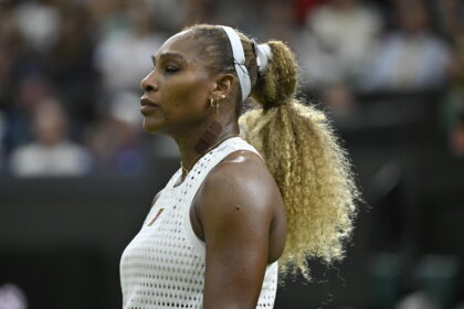 Profile view of a female tennis player wearing a white perforated sleeveless top and a white headband during a match, with a blurred crowd in the background.