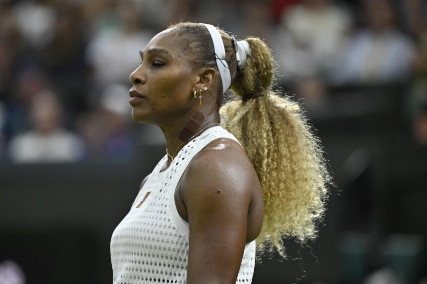 Profile view of a female tennis player wearing a white perforated sleeveless top and a white headband during a match, with a blurred crowd in the background.