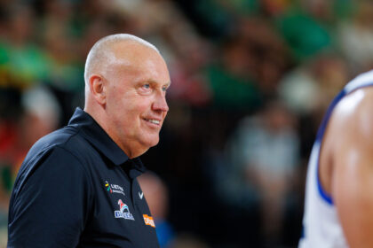 Smiling bald man in a navy polo, likely a basketball coach, on a court with a blurred crowd in the background.