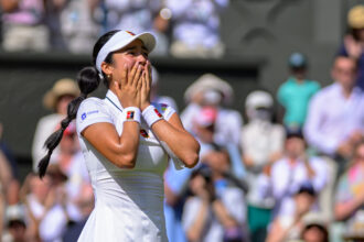 Female tennis player in a white outfit and visor covers her mouth with her hands in celebration on a tennis court, with a blurred crowd behind her.