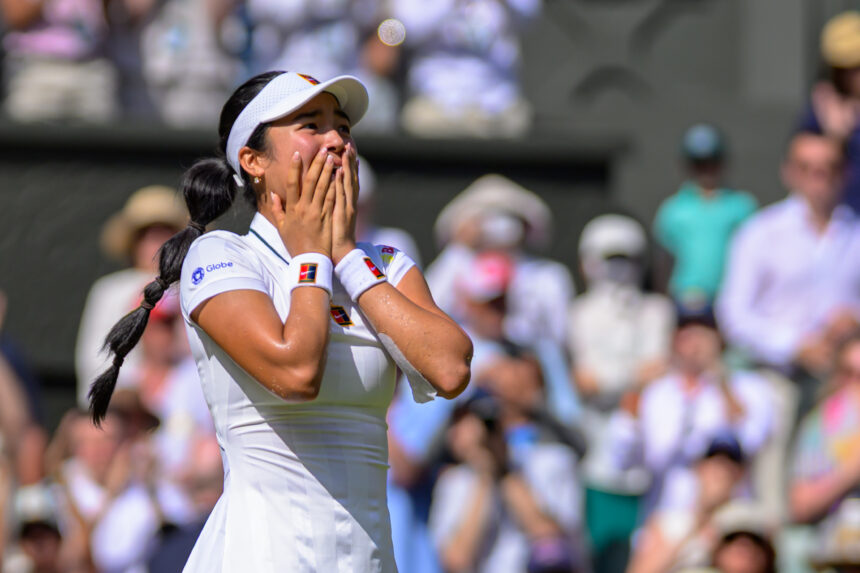 Female tennis player in a white outfit and visor covers her mouth with her hands in celebration on a tennis court, with a blurred crowd behind her.