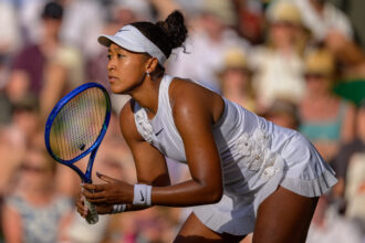 Female tennis player in a white outfit and visor, leaning forward with a blue racket as she prepares to hit the ball, crowd blurred in the background.