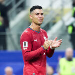 Soccer player in a red jersey applauds on the field, with blurred stadium staff in neon vests in the background.