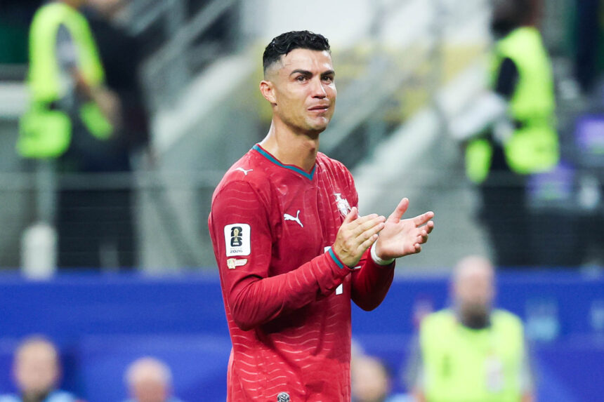 Soccer player in a red jersey applauds on the field, with blurred stadium staff in neon vests in the background.
